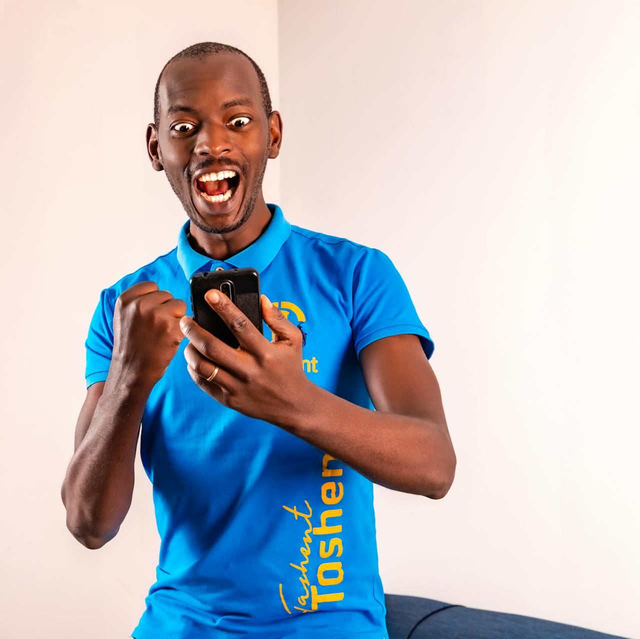 A cheerful man in a blue polo shirt expressing excitement while looking at a smartphone indoors.